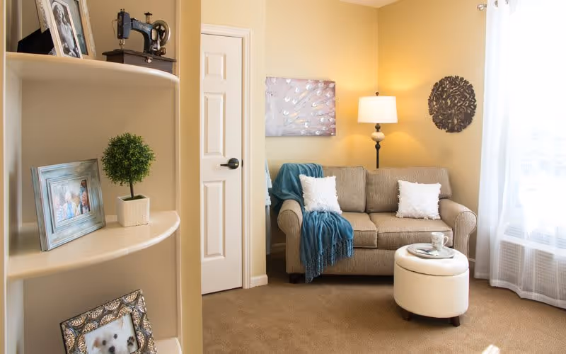 Cozy living room with a beige loveseat, throw blanket and pillows, round ottoman, a floor lamp, shelving with framed photos, and sheer curtains.