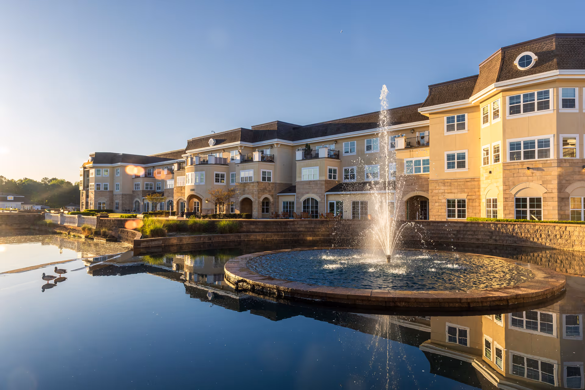 Exterior view of a large, multi-story retirement community building with beige and stone facade, multiple windows, and balconies. In front of the building is a circular water fountain in a pond with clear reflections of the building and fountain. There are also a few ducks swimming in the pond. The sky is clear and the scene is lit by warm sunlight.