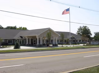 Exterior view of a single-story building with a covered entrance and an American flag flying on a flagpole in front. The building is surrounded by a lawn and trees, with a sign that reads Sugar Creek Alzheimer's Special Care Center near the entrance.