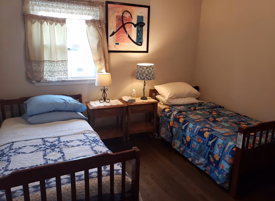 A small bedroom with two single wooden beds, each with different bedding. The left bed has a white and blue quilt with blue pillows, and the right bed has a colorful patterned blanket with white pillows. Between the beds are two wooden nightstands with lace doilies, a lamp with a patterned shade, a smaller lamp, and a bottle of lotion. A window with lace curtains is on the left wall, and a framed abstract painting hangs above the nightstands. The room has wooden flooring and beige walls.