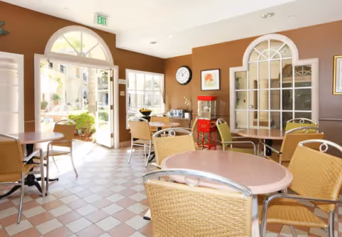 A bright dining area with round tables and wicker chairs. The room has large arched windows and a door leading outside to a garden area. There is a clock on the wall, a popcorn machine, and framed artwork. The floor is tiled with a checkered pattern in light and dark tones.