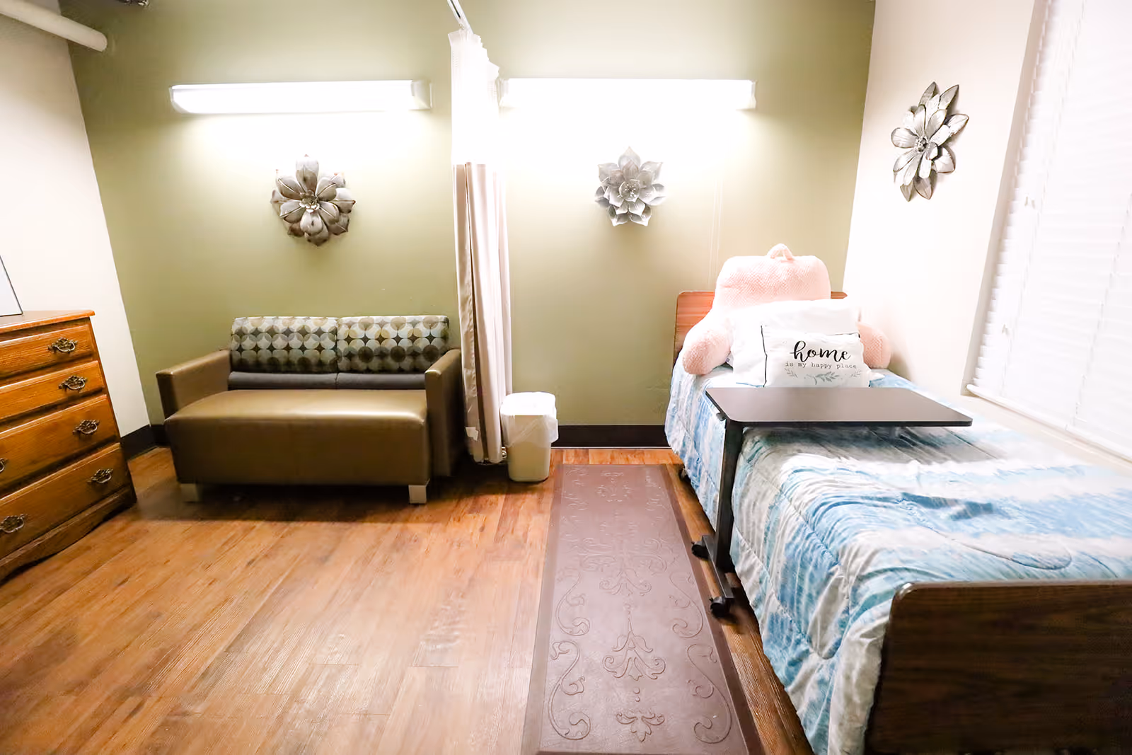 A cozy room with a single bed on the right side, covered with a blue and white blanket, a large pink teddy bear, and a white pillow with the word 'home' on it. Next to the bed is a small overbed table. On the left side of the room is a brown loveseat with patterned cushions, a wooden dresser with drawers, and a trash bin. The walls are painted light green and beige, decorated with three metal flower wall hangings. The floor is wooden with a decorative runner rug in the center.