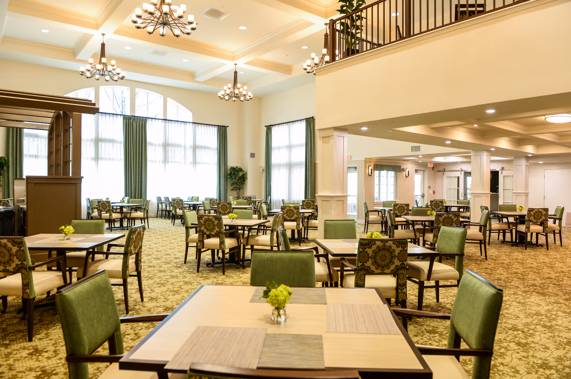 A spacious and well-lit dining room with multiple tables and chairs arranged neatly. The room features large windows with green curtains, chandeliers hanging from a coffered ceiling, and small floral centerpieces on each table. The carpet has a patterned design, and there is a balcony or mezzanine level visible above.
