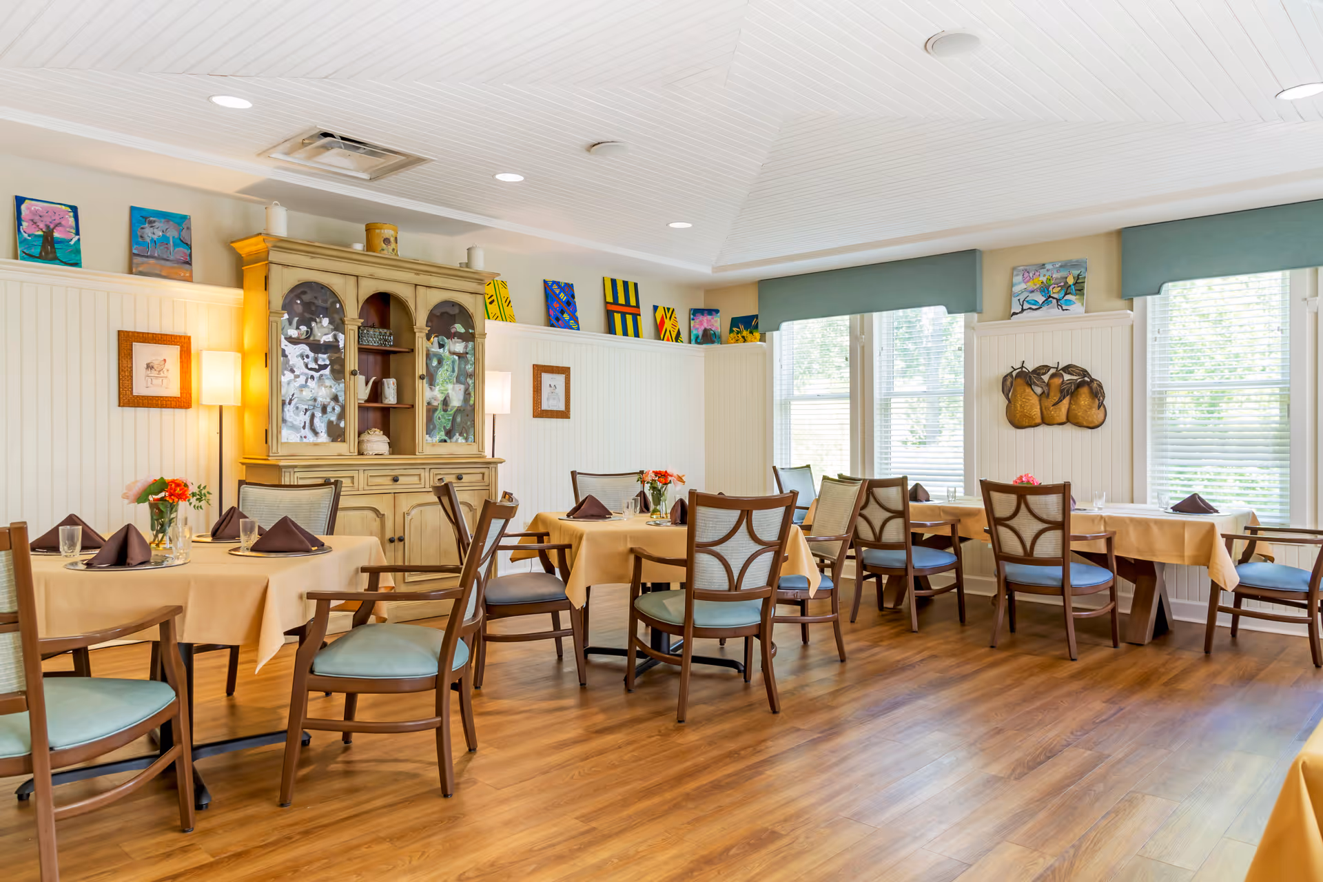 Bright, well-lit dining room with multiple tables set with napkins and chairs, a wooden cabinet, artwork, and large windows.