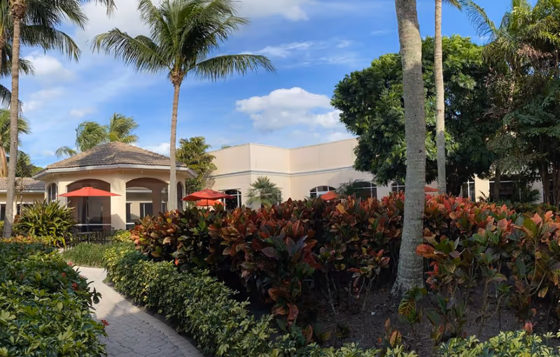 Outdoor garden area at Courtyard Gardens of Jupiter featuring a paved walkway surrounded by lush green and red foliage, palm trees, and a building with a covered patio and red umbrellas under a partly cloudy blue sky.