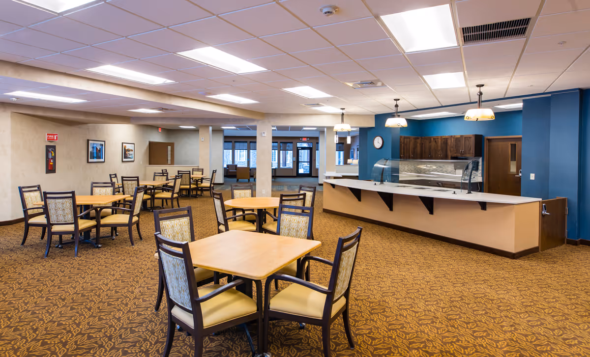 A spacious dining area in a senior living facility with multiple wooden tables and chairs arranged on a patterned carpet. The room features a serving counter with a glass sneeze guard, pendant lights hanging from the ceiling, and a blue accent wall with wooden cabinets and a clock. The background shows an open doorway leading to another seating area with large windows.