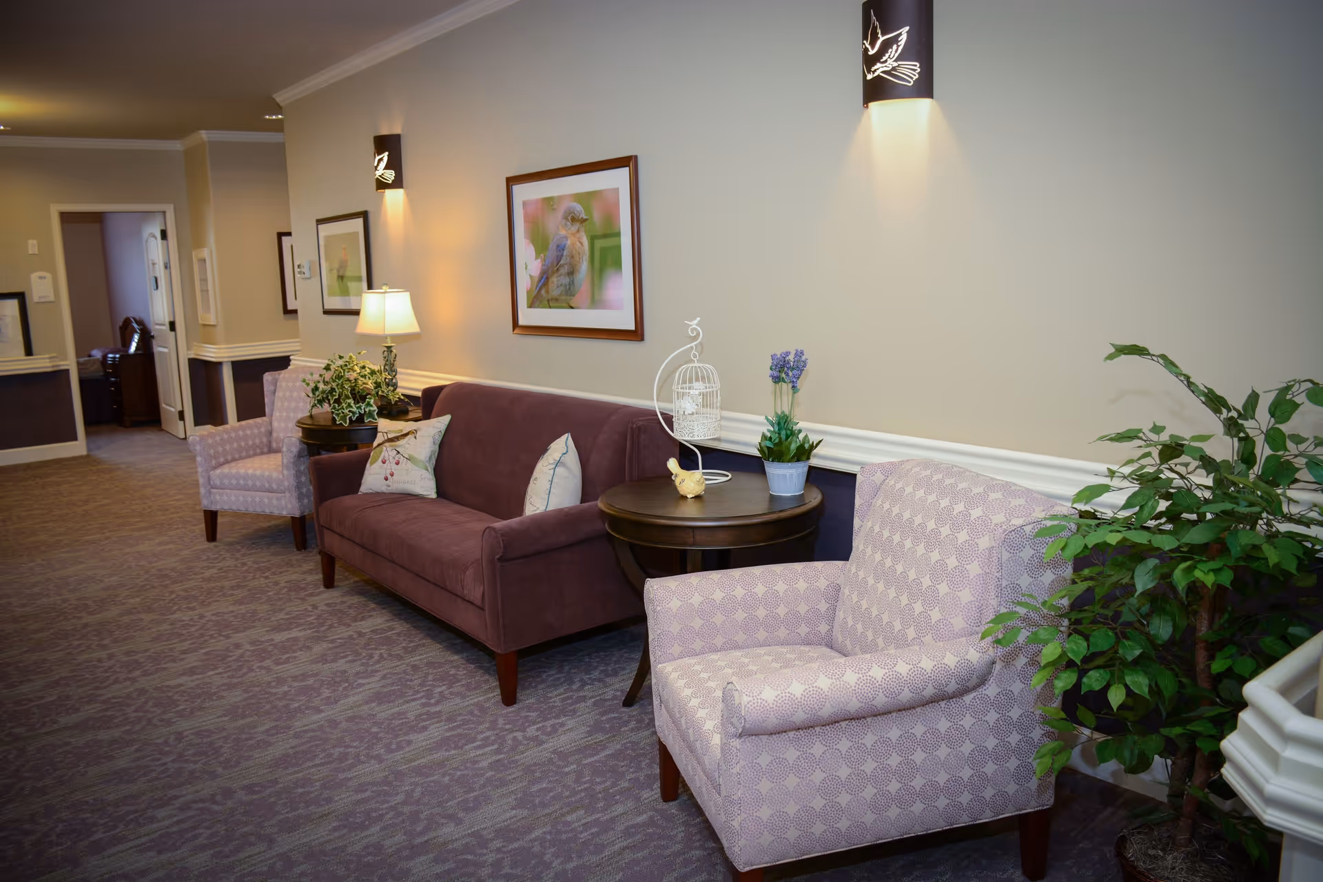 A cozy seating area in a hallway with a purple sofa, two patterned armchairs, a round wooden side table with decorative items, a table lamp, framed bird artwork on the wall, and a potted plant in the corner.