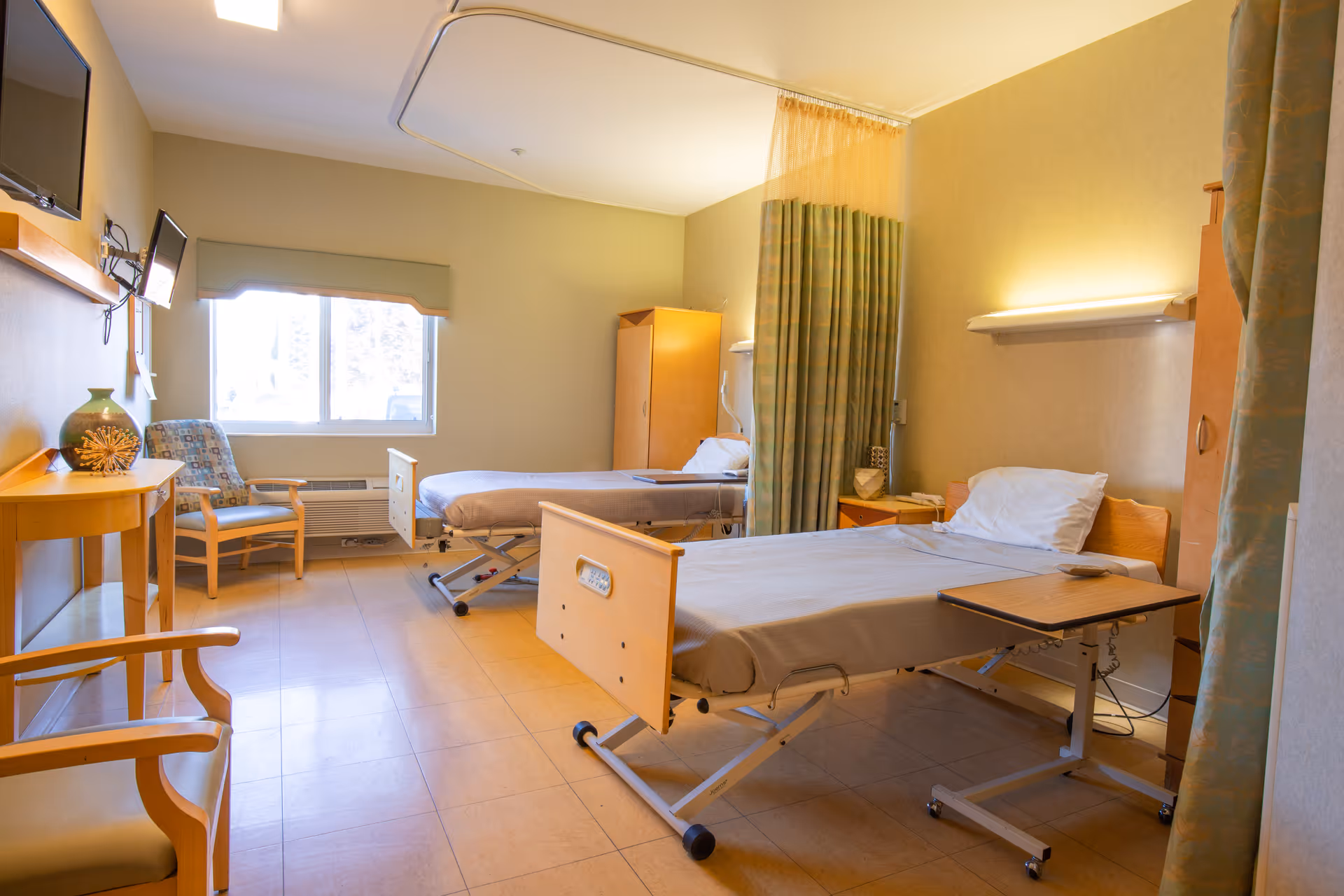 A bright room with two hospital-style beds separated by a green privacy curtain. Each bed has a pillow and a small overbed table. There is a wooden chair and a small table with a decorative vase near a window with a green valance. The walls are light-colored, and there is a wall-mounted TV on the left side.