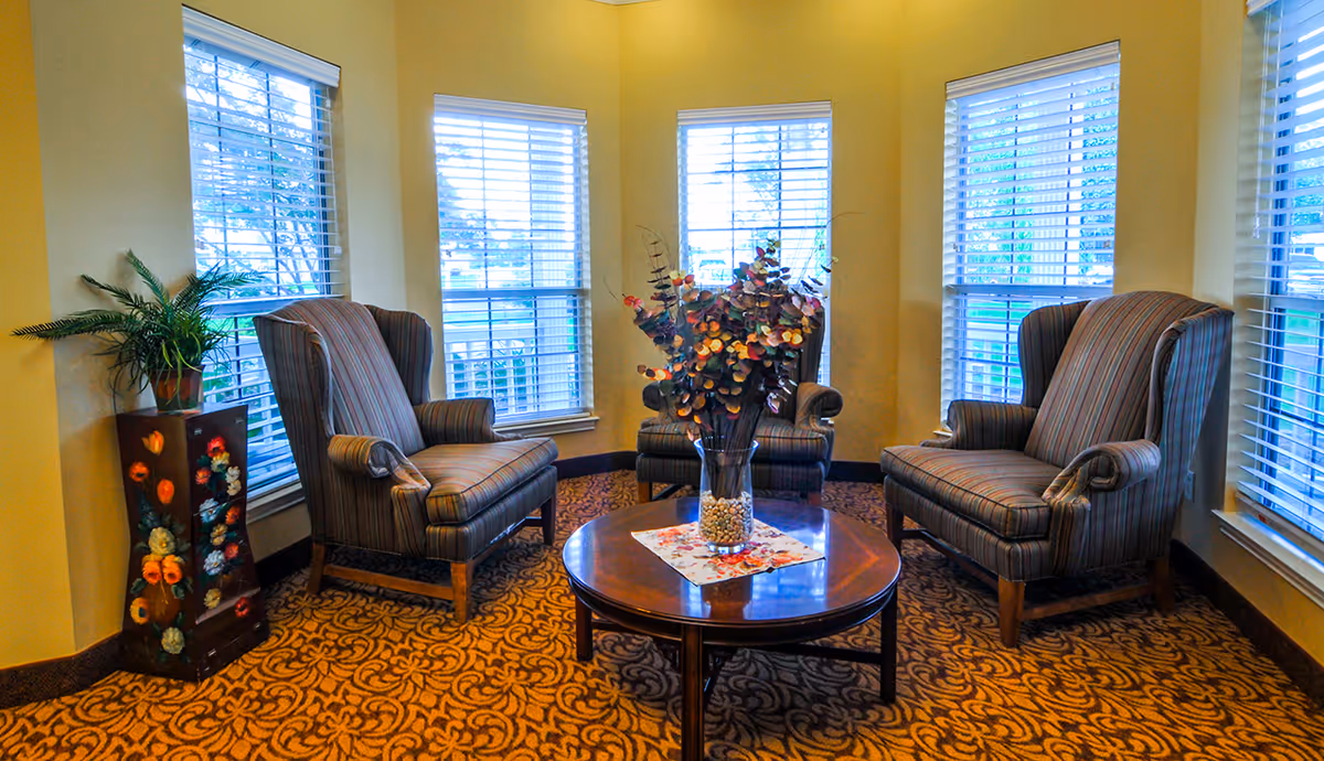 A cozy sitting area with three striped armchairs arranged around a round wooden coffee table that has a glass vase with decorative foliage. The room has yellow walls, patterned carpet, and multiple windows with white blinds letting in natural light. A small decorative stand with a potted plant is placed in the corner.