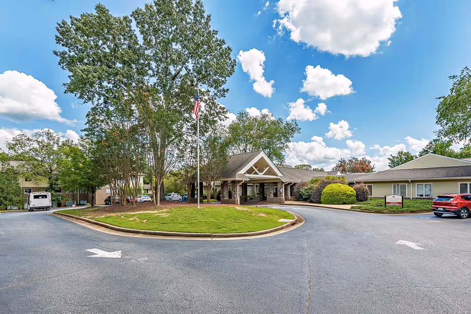 Front entrance of a senior living facility with a circular driveway, flagpole, landscaping and parked cars under a blue sky.