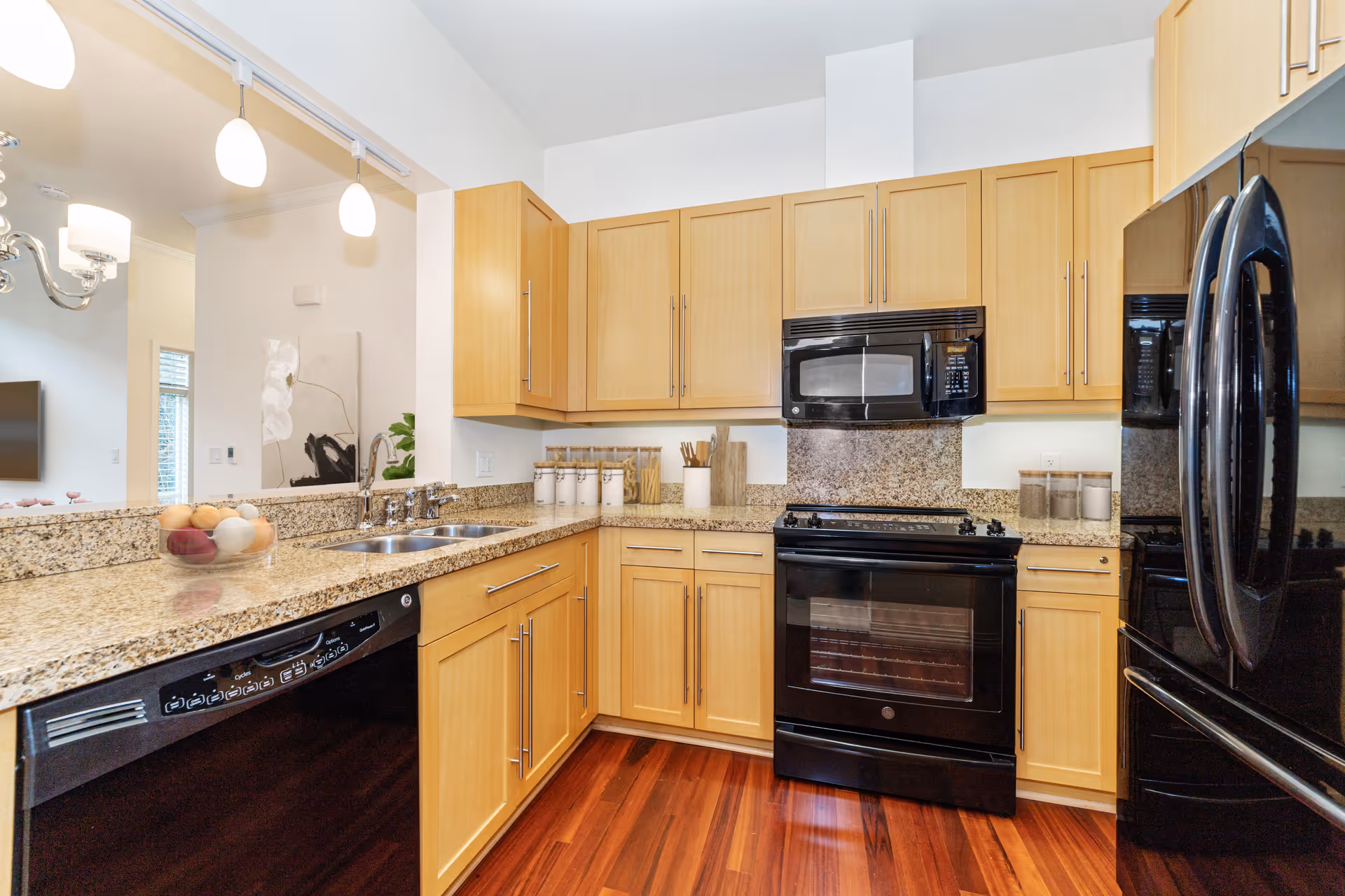 Modern kitchen with light wood cabinets, granite countertops, black appliances including a refrigerator, oven, microwave, and dishwasher. The kitchen has a double sink and wooden flooring. Pendant lights hang above the counter, and a glimpse of a living area with a TV and wall art is visible in the background.