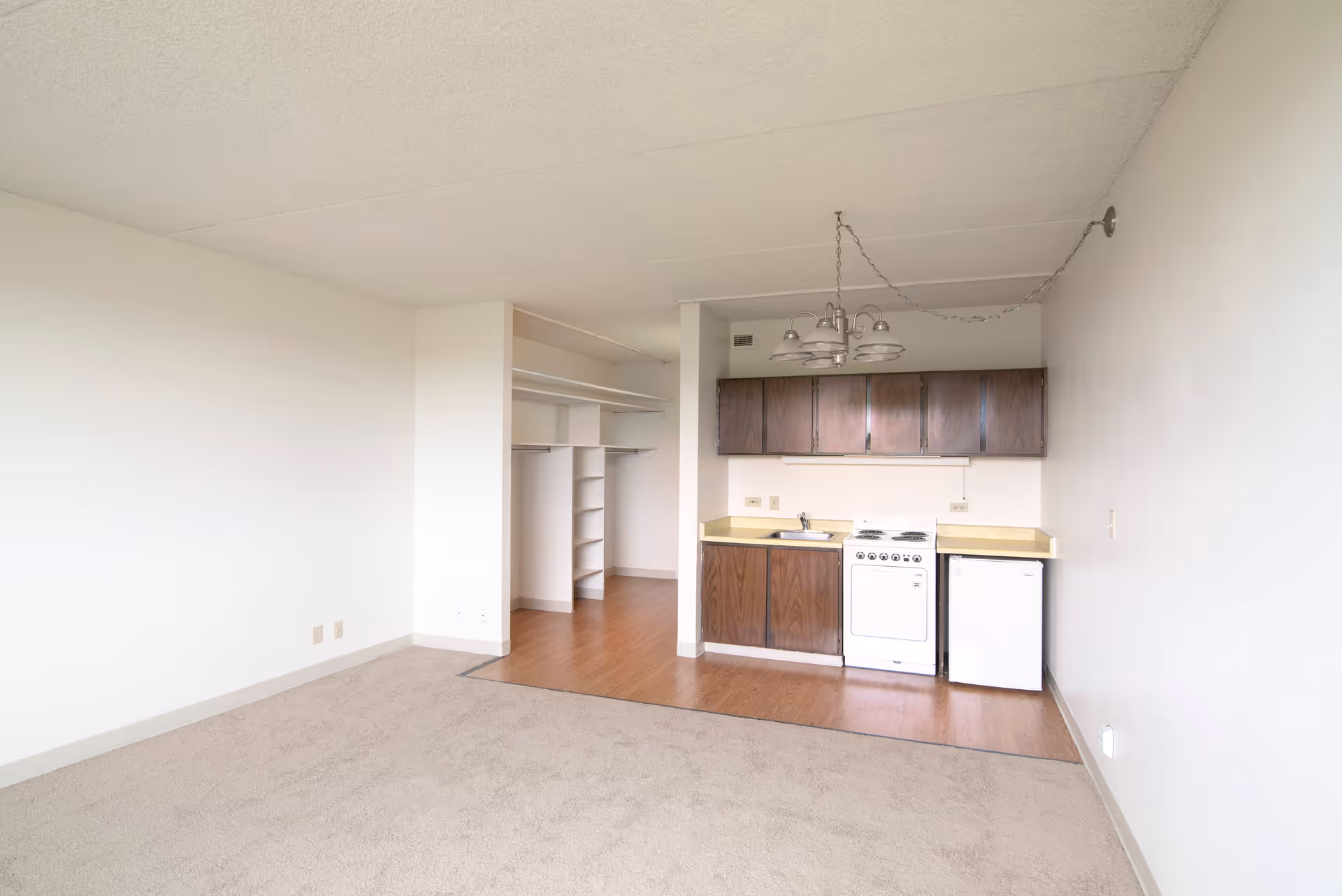 Studio apartment interior with a small kitchenette, open closet shelving, and a carpeted living area.