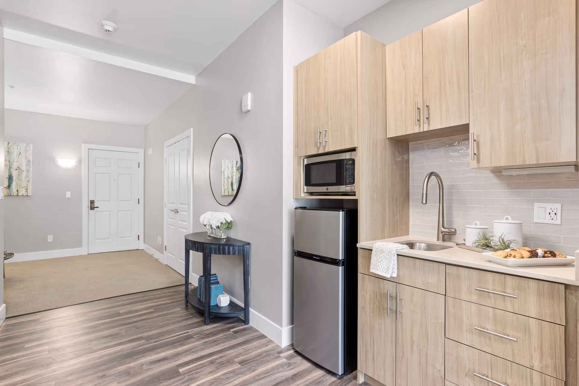 Interior view of a modern kitchenette area with light wood cabinets, a small stainless steel refrigerator, a microwave, a sink with a faucet, and a countertop with a towel and a tray of pastries. Adjacent to the kitchenette is a hallway with a round mirror above a small black table holding a vase of white flowers and some books. The hallway leads to a white door with a silver handle and a wall-mounted light fixture.