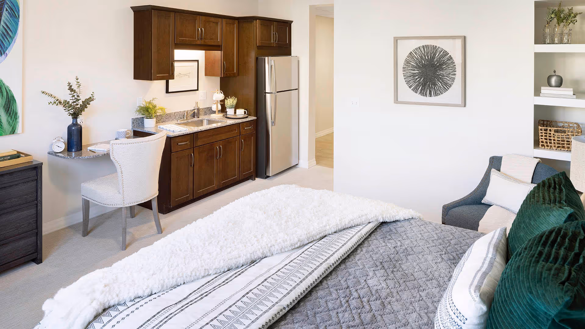 A cozy studio apartment featuring a bed with gray and white bedding and green pillows, a small kitchenette with dark wood cabinets, a stainless steel refrigerator, a sink, and a small desk with a white upholstered chair. The room also includes a gray armchair, wall art, and built-in shelves with decorative items.