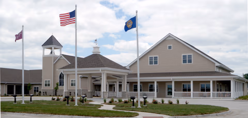 Exterior view of Brookestone Acres facility showing a large beige building with a covered entrance, three flagpoles with flags including the American flag, and a well-maintained lawn and walkway in front.
