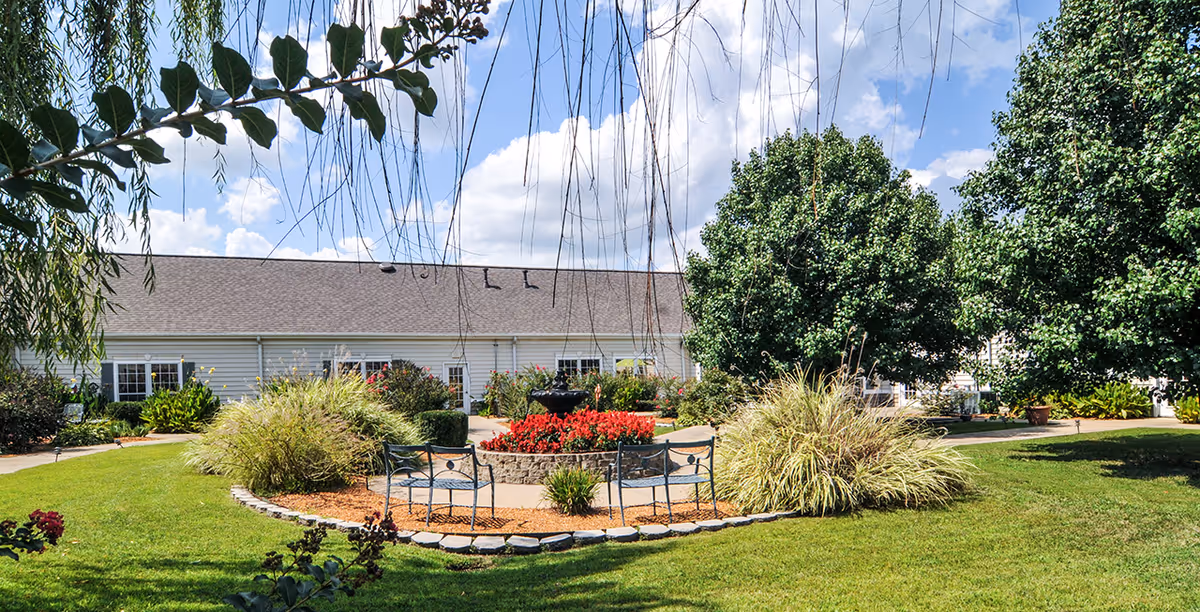 Sunny landscaped courtyard with benches, a central flowerbed and fountain in front of a single-story building.