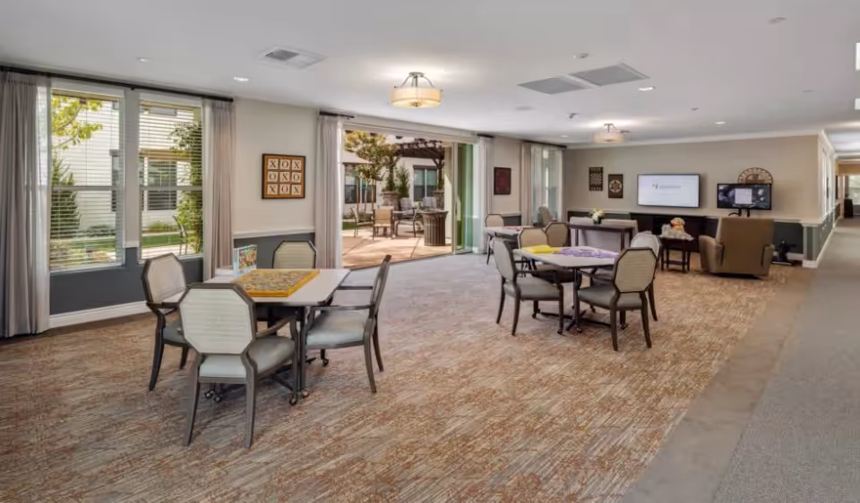 A spacious common area in a senior living facility with multiple tables and chairs arranged for social activities. Large windows and glass doors open to an outdoor patio with seating and greenery. The room is carpeted and well-lit with ceiling lights. A television and comfortable armchairs are visible in the background.