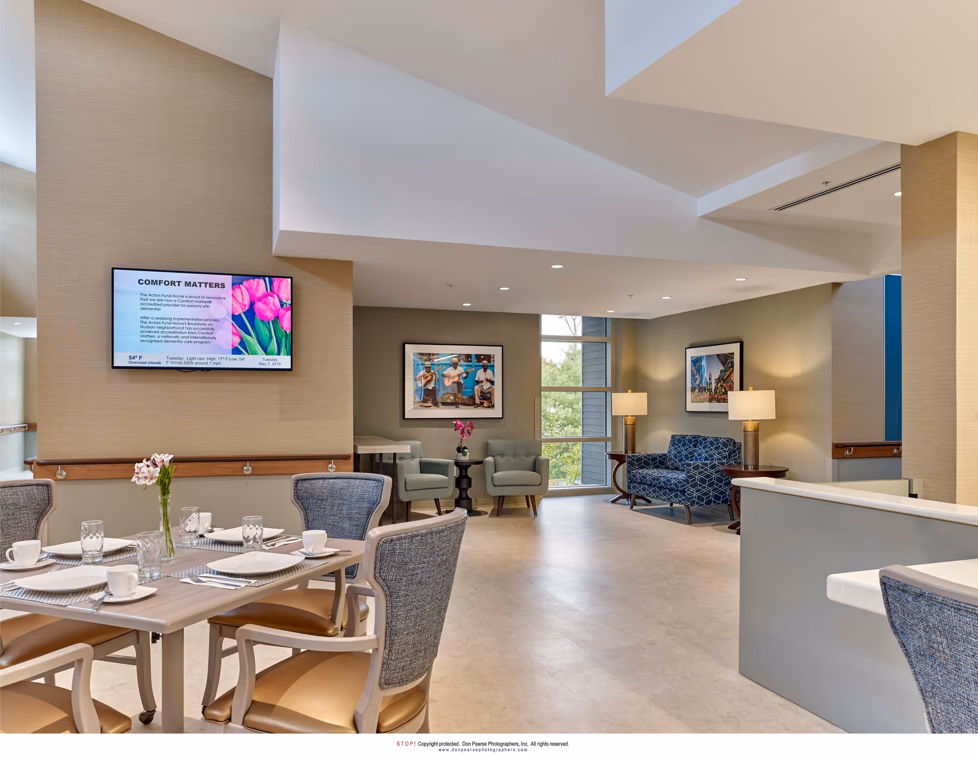 A bright and spacious common area in a senior living facility featuring a dining table set with plates, cups, and glasses. The room has comfortable seating including armchairs and a patterned loveseat, with framed artwork on the walls and two table lamps providing warm lighting. A flat-screen TV mounted on the wall displays information titled 'Comfort Matters'. Large windows allow natural light to fill the space.