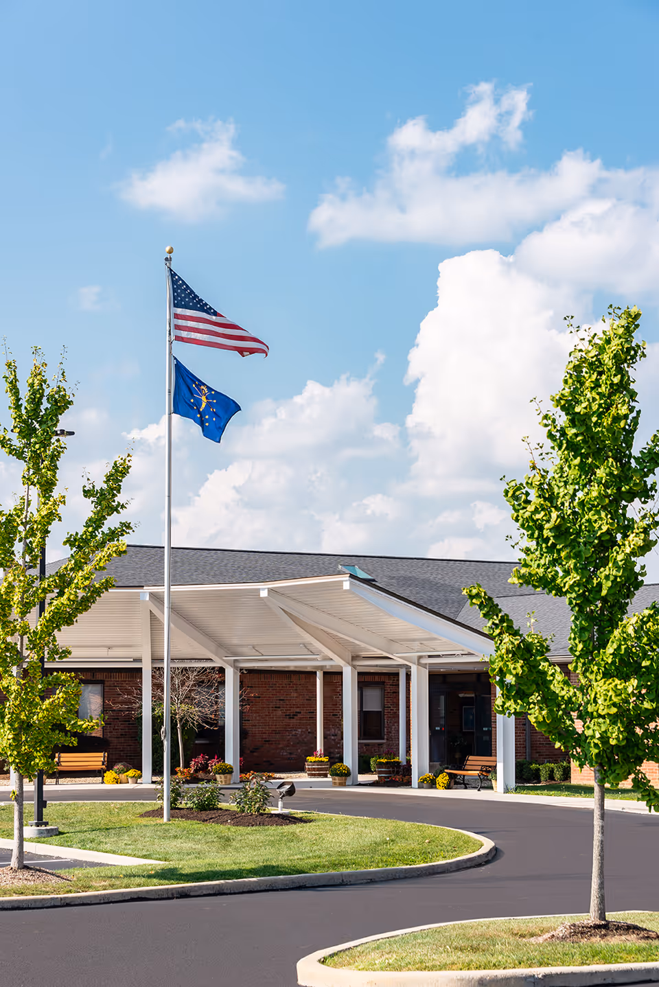Entrance of a brick building with a covered drop-off area, two flagpoles displaying the American flag and a blue state flag, surrounded by green trees and a curved driveway under a partly cloudy blue sky.