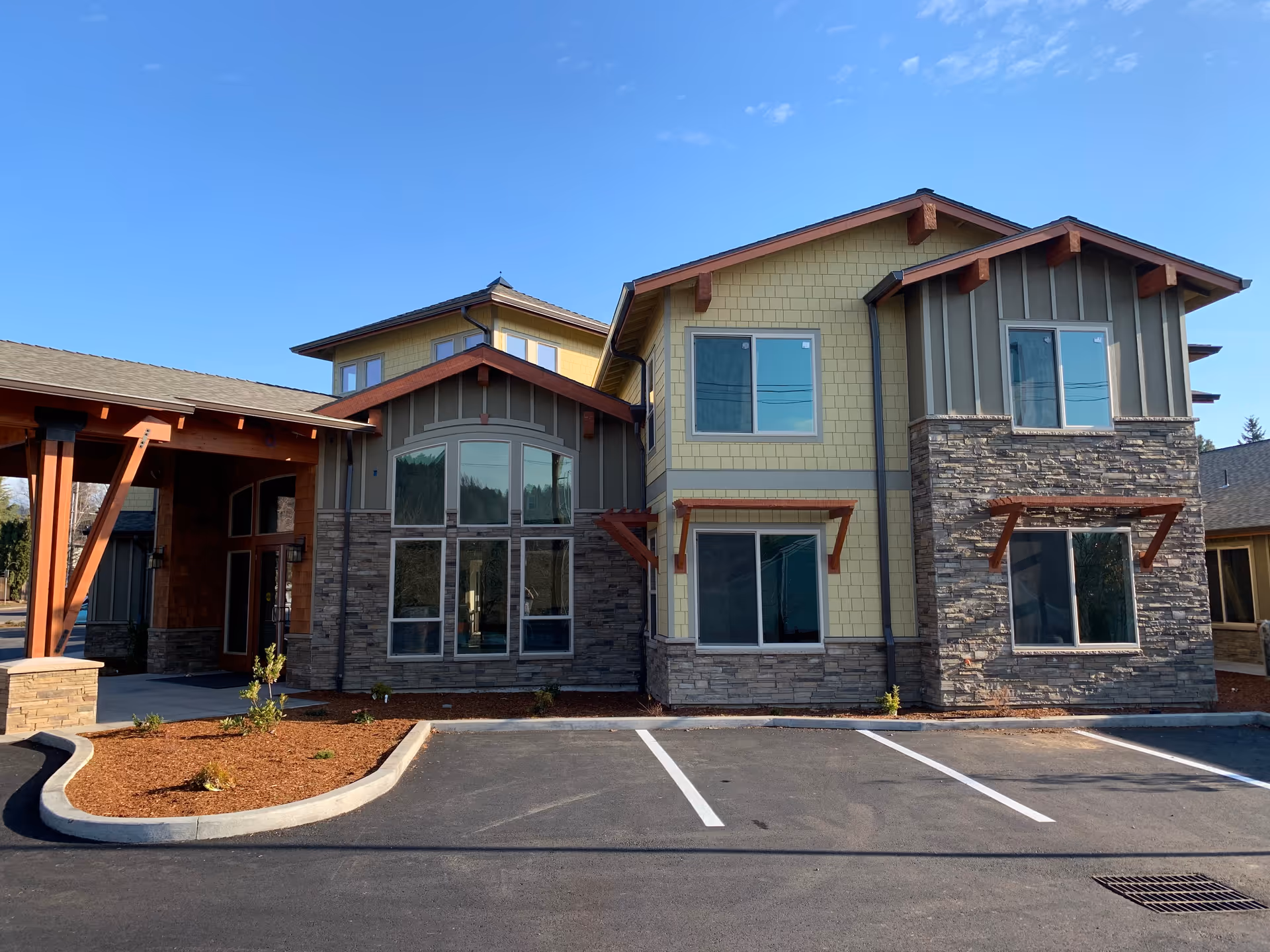 Front exterior of a two-story memory care building with stone and yellow siding, a covered entryway, and empty parking spaces.