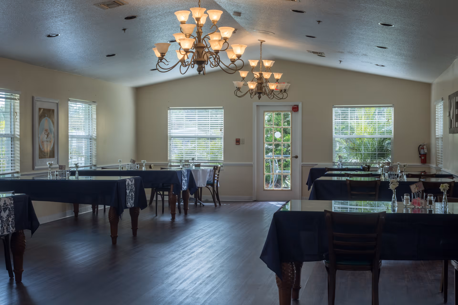 Dining room with several tables covered in dark tablecloths, chandeliers, windows, and a glass door leading outside.
