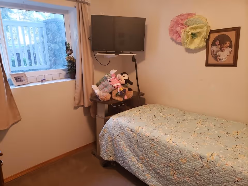 Small furnished bedroom with a single bed, wall-mounted TV, stuffed animals on a bedside table, and a window.