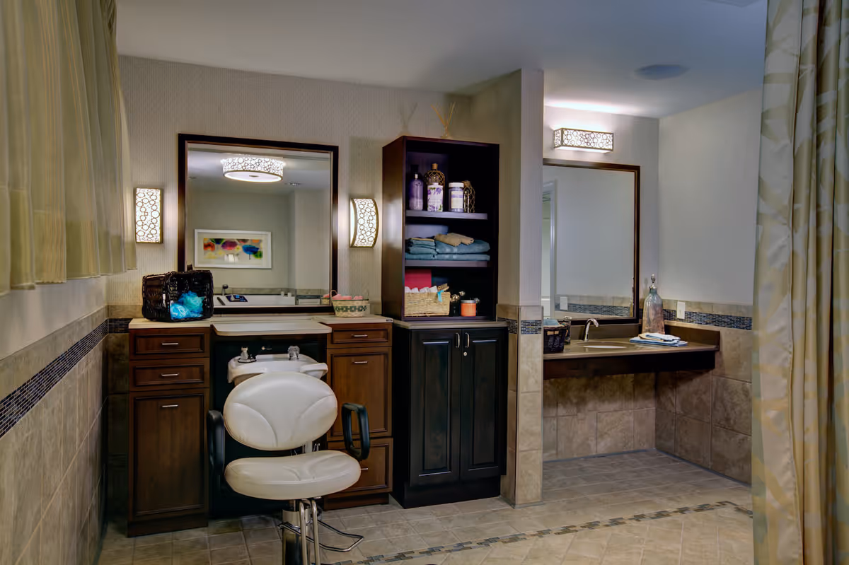 Interior view of a well-lit salon area in a senior living facility with a white salon chair in front of a sink and mirror, wooden cabinets with towels and toiletries, and a separate vanity area with a sink and mirror. The room has tiled floors and walls with decorative lighting fixtures.