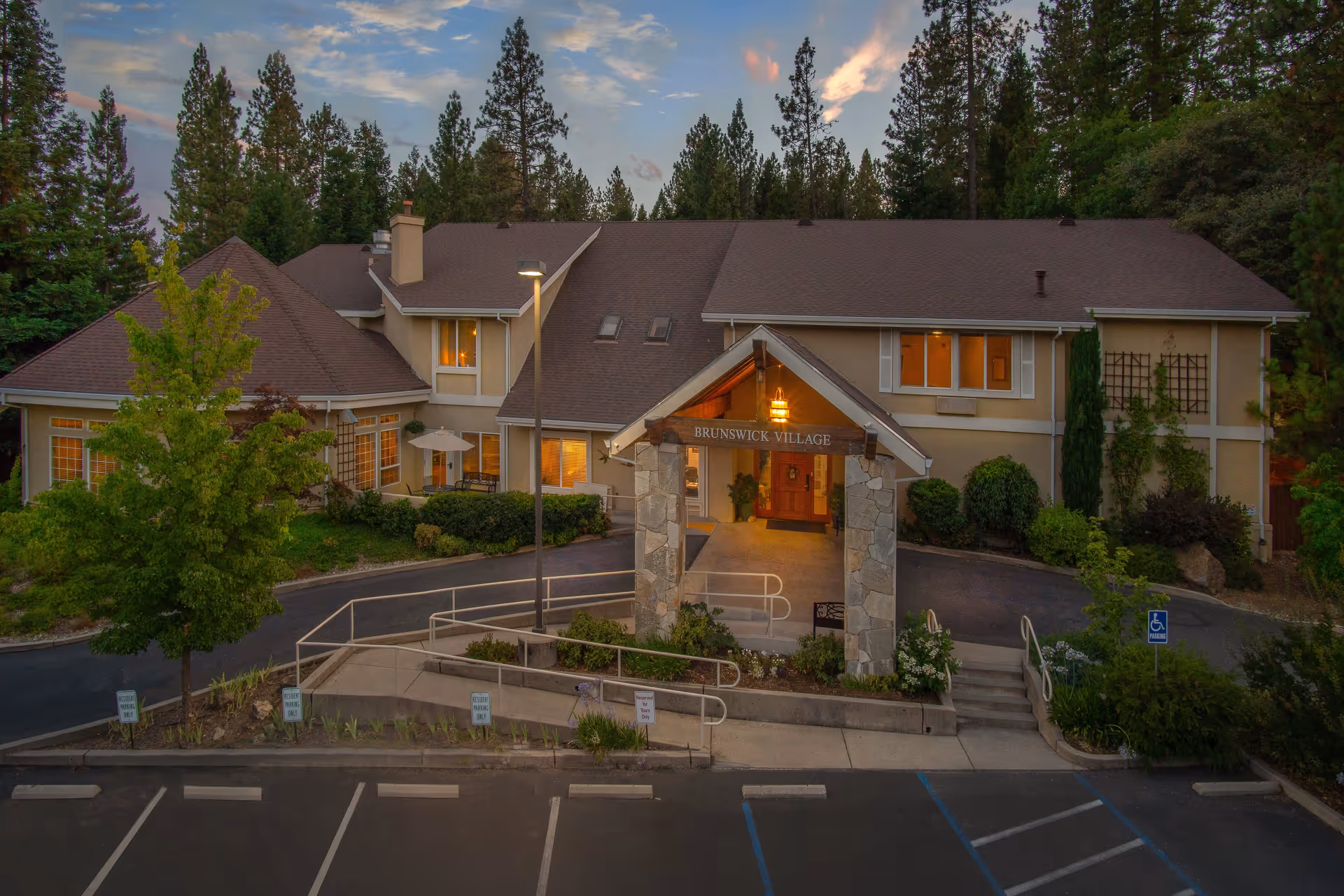 Exterior view of Brunswick Village Assisted Living facility at dusk, showing a two-story building surrounded by trees and landscaping. The entrance features a covered stone archway with a hanging light and the facility name displayed. There is a parking area with marked spaces and a handicap parking sign in front.