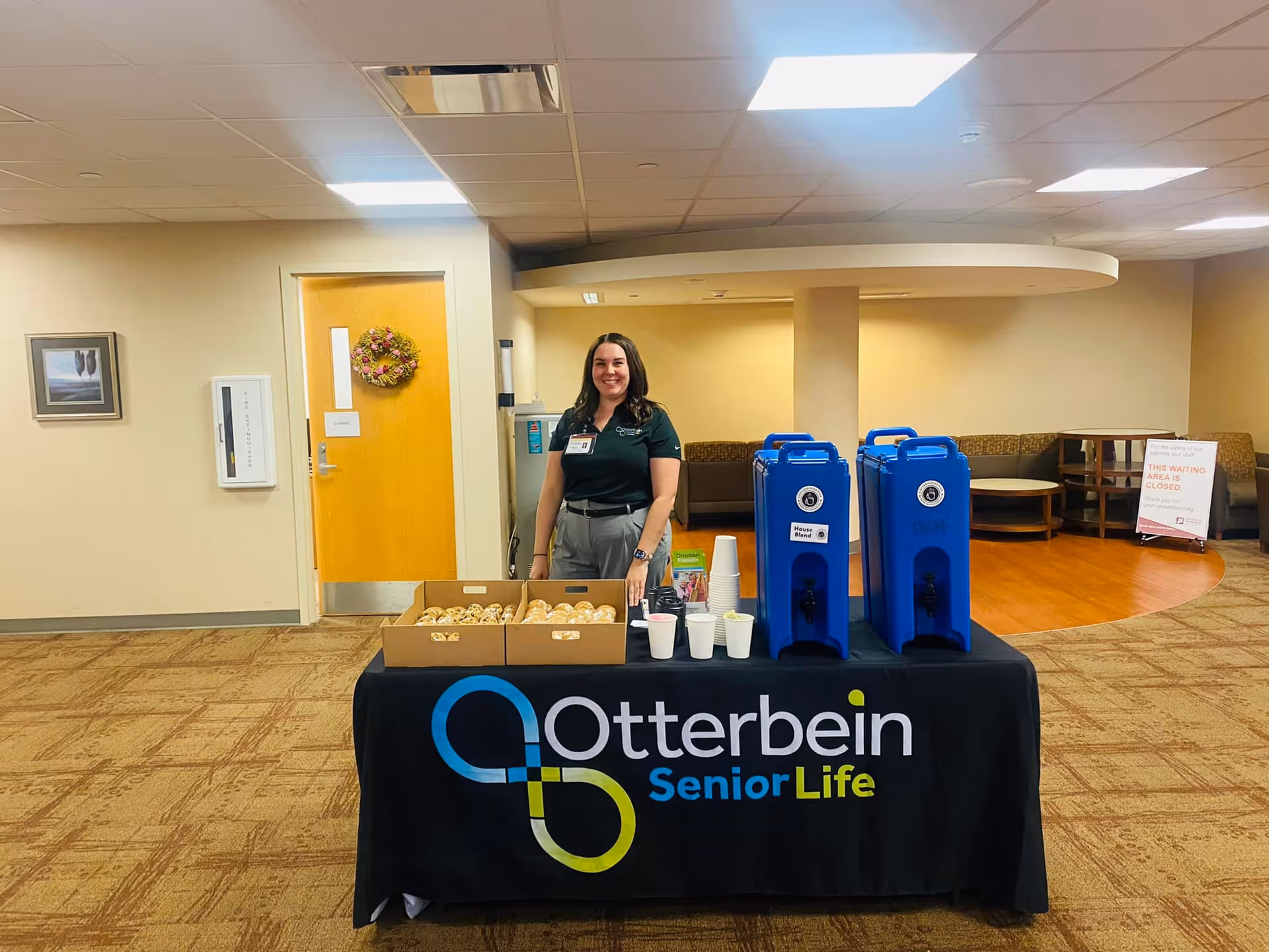 A woman stands behind a table covered with a black tablecloth that has the Otterbein SeniorLife logo. On the table are two large blue beverage dispensers, several white cups, and boxes filled with bagels. The setting appears to be an indoor common area with carpeted flooring, beige walls, and a wooden door decorated with a wreath. There is seating in the background and a sign indicating a waiting area is closed.