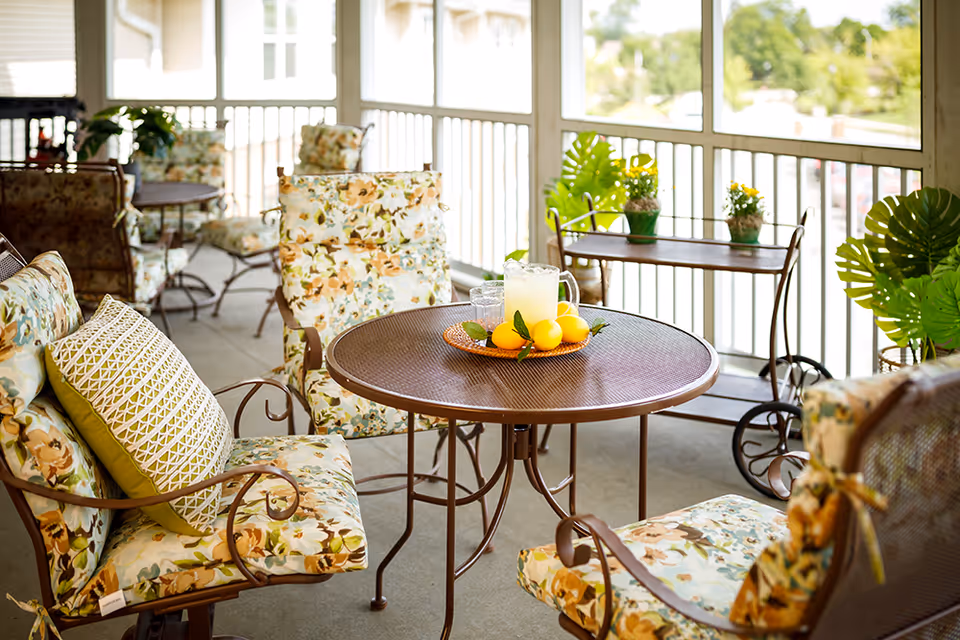 A cozy outdoor patio area with floral cushioned chairs around a round metal table. On the table, there is a pitcher of lemonade and a plate with lemons. The patio is enclosed with white railings and decorated with green plants and small potted flowers on a serving cart in the background.