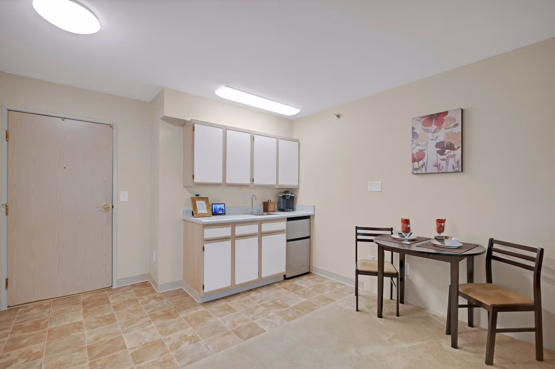 Small studio-style kitchenette with cabinets, sink, mini-fridge and a two-person dining table against a neutral wall.