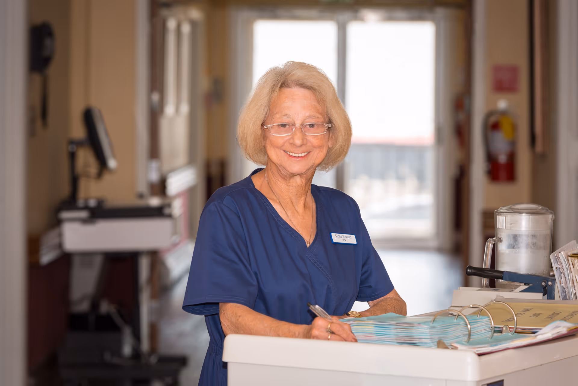 A smiling elderly woman wearing glasses and a navy blue uniform stands behind a desk or counter in a hallway, holding a pen and looking at an open binder with documents. The background shows a bright doorway and some medical equipment.