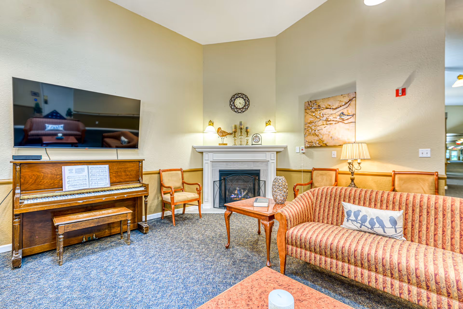 A cozy living room area in Creekside Place Memory Care featuring a striped sofa with a bird-patterned pillow, a wooden coffee table with a decorative item and book, two wooden chairs with beige cushions, a fireplace with candles and a clock above it, a wall-mounted flat-screen TV, and an upright piano with sheet music. The room has beige walls, carpeted floor, and warm lighting.