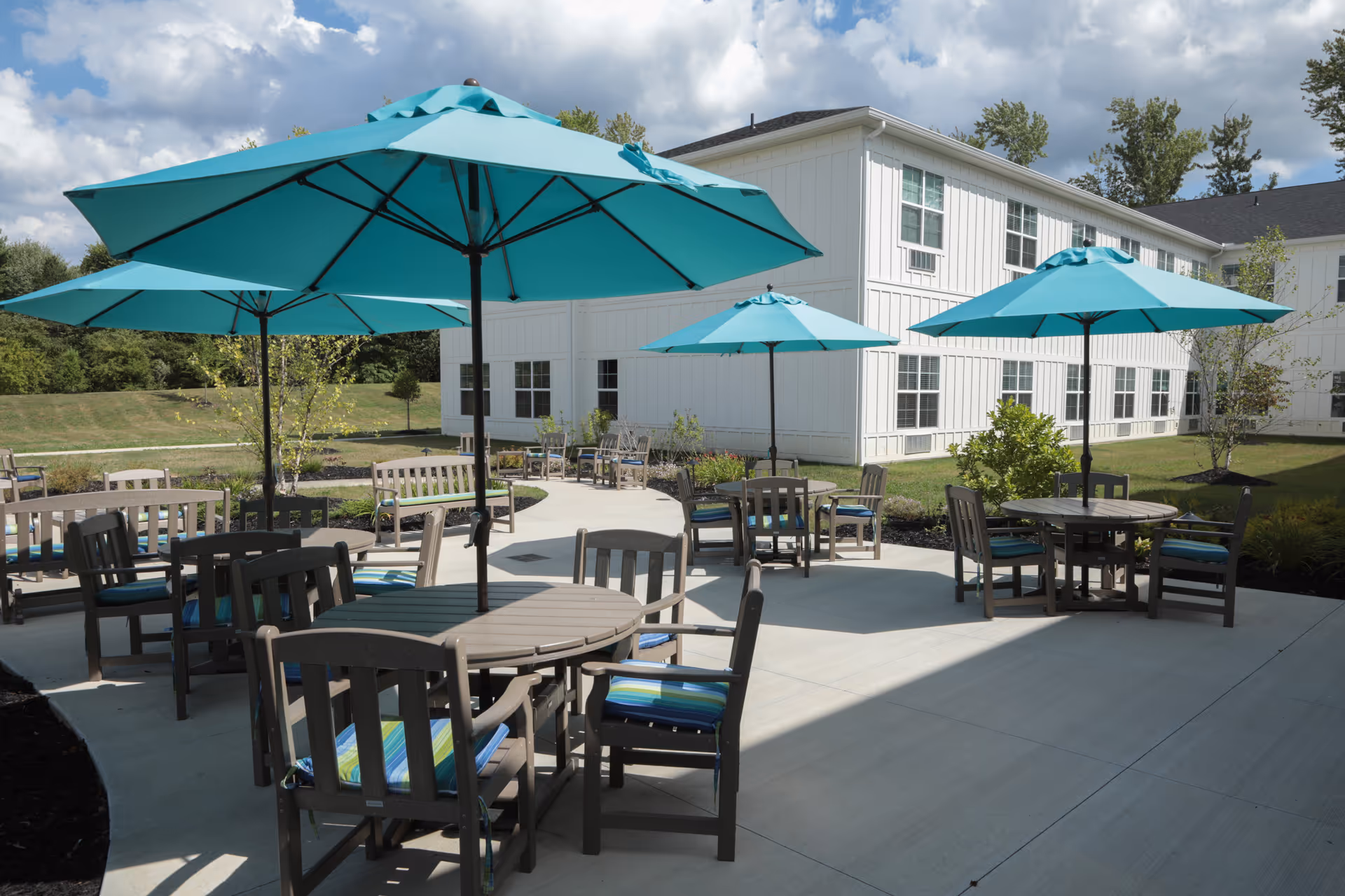 Outdoor patio area at Heritage Crossing with multiple round tables and chairs, each shaded by large turquoise umbrellas. The patio is surrounded by a well-maintained lawn and landscaping, with a white two-story building in the background under a partly cloudy sky.