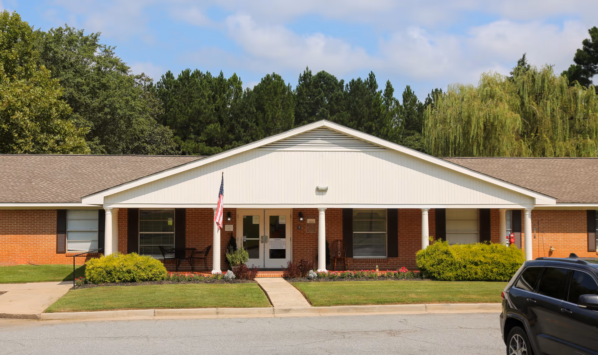 Front exterior view of a single-story brick building with a white gabled roof and columns at the entrance. There is an American flag near the door, some bushes and flowers in front, and a black SUV parked on the right side. Trees and a blue sky with clouds are visible in the background.