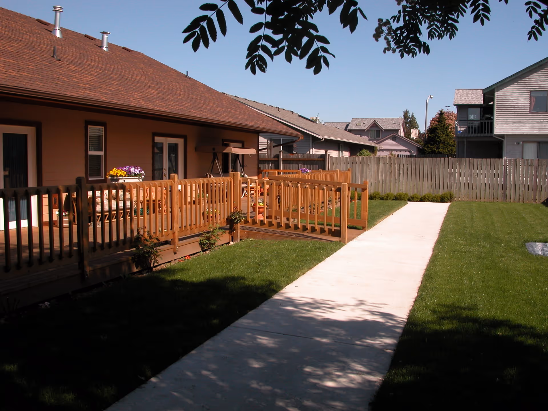 Outdoor view of a senior living facility with a wooden deck and railing, a concrete walkway, green grass lawn, and a wooden fence in the background under a clear blue sky.