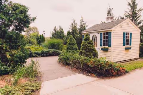 Small beige building with green shutters sits among landscaped shrubs beside a paved walkway.