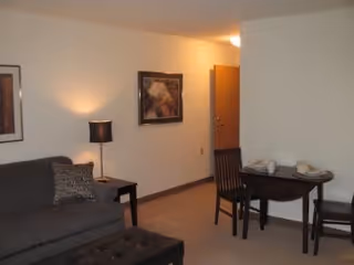 Small furnished living room with a sofa, side table and lamp, wall art, a small dining table with two chairs, and an entry door.