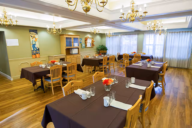 A dining room with several wooden tables covered with dark brown tablecloths, each set with white napkins, cups, glasses, and small flower arrangements. The room has wooden floors, light green walls, multiple chandeliers, large windows with sheer curtains, and a wooden cabinet against one wall.