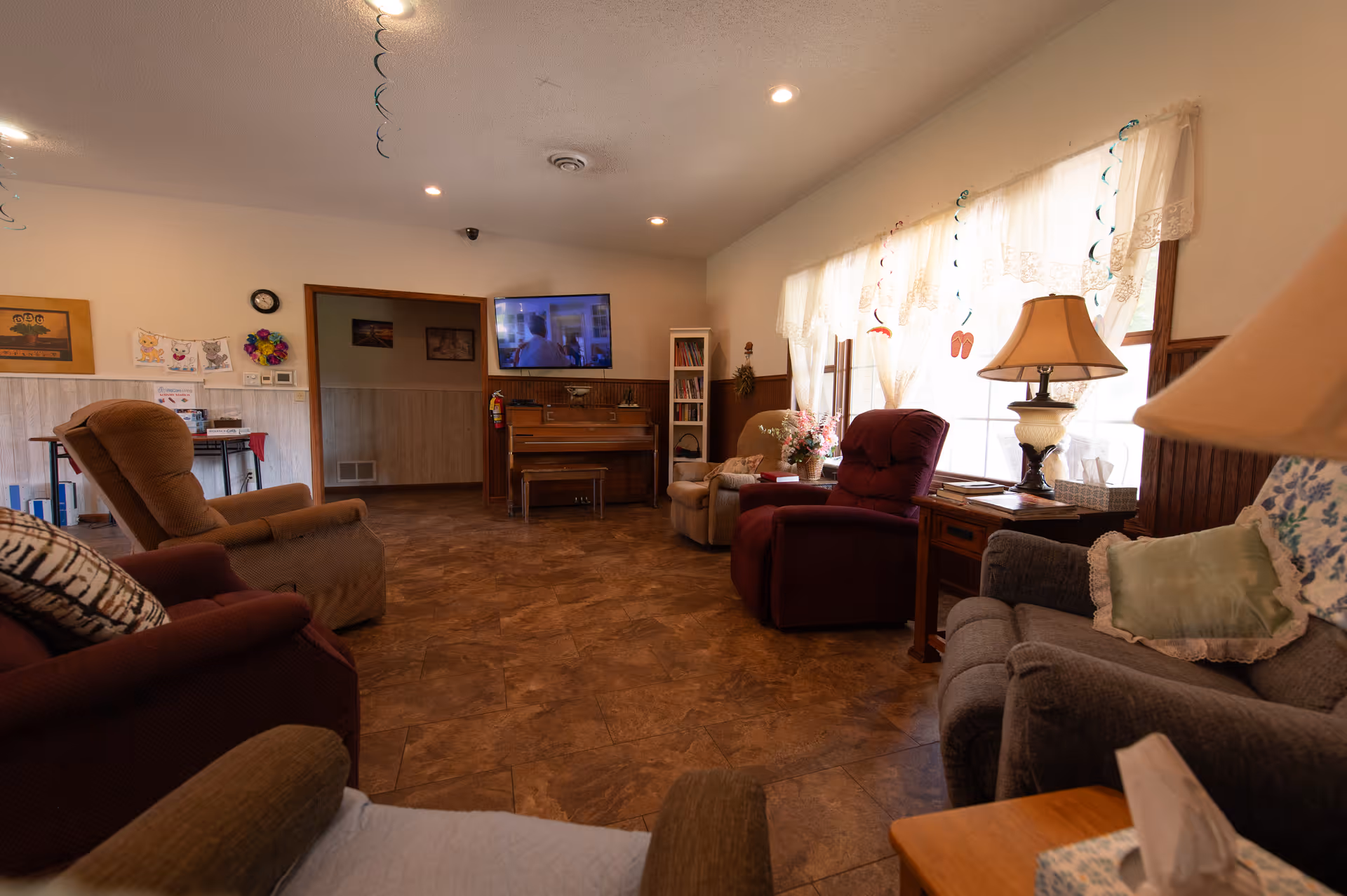 Sunlit senior living common room with multiple recliners, side tables, a piano and a wall-mounted TV.