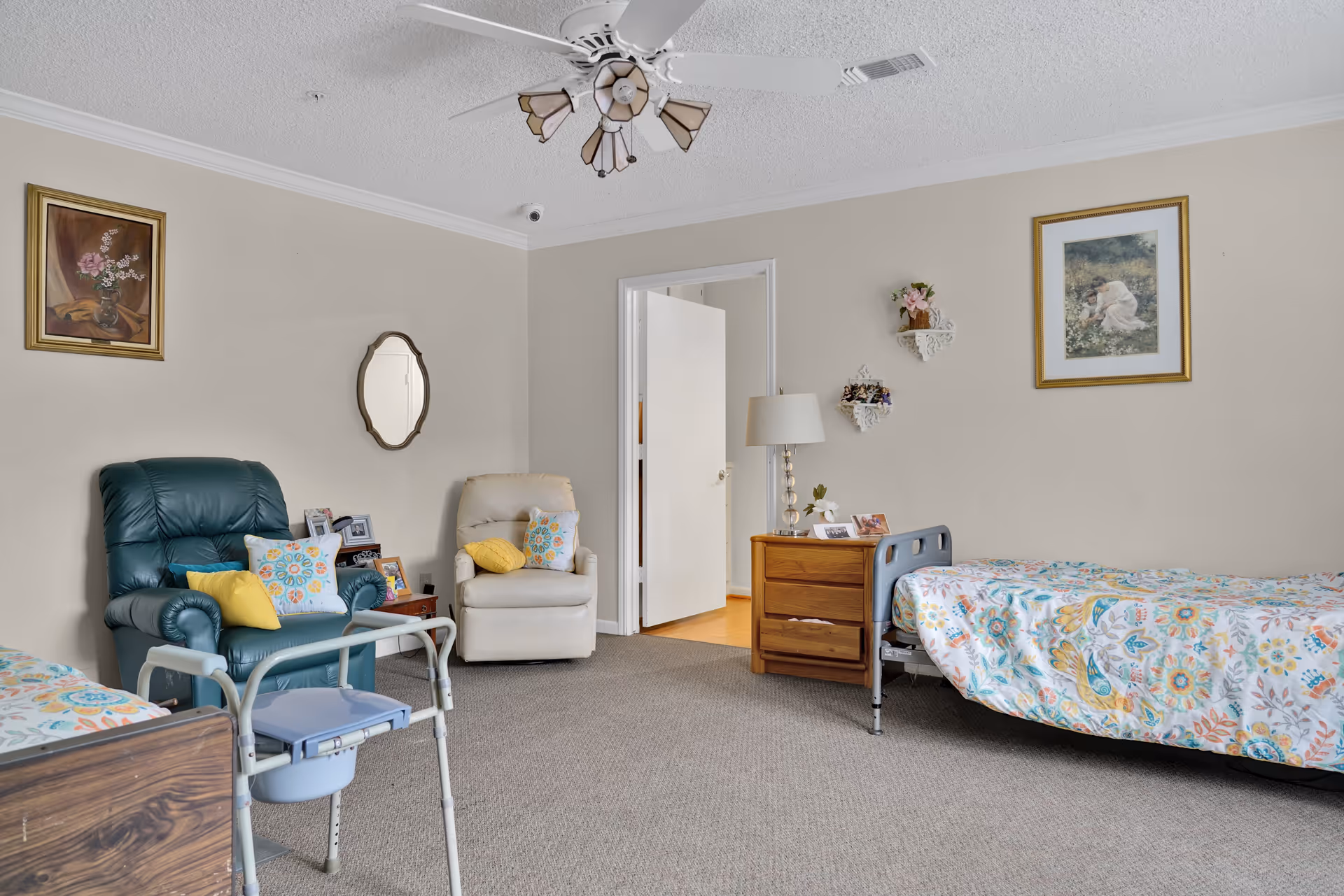 A cozy bedroom in a senior living facility with two beds covered in colorful patterned bedspreads. There are two armchairs with decorative pillows, a wooden nightstand with a lamp and framed photos, and wall decorations including framed paintings and small shelves with flowers. A ceiling fan with lights is mounted on the ceiling, and a walker is positioned near one of the beds.