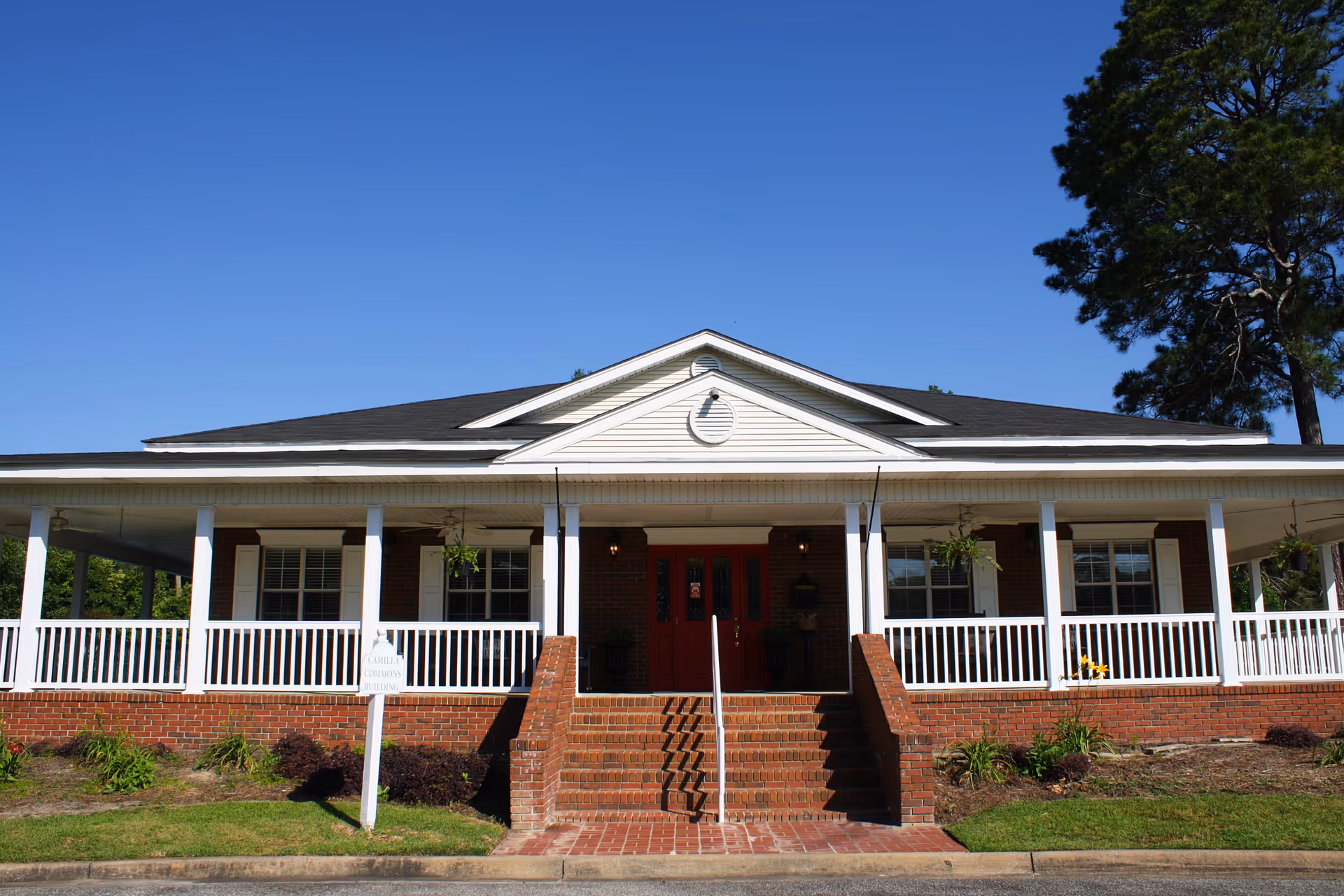 Front exterior view of a single-story brick building with a white railing porch, red double doors, and a small set of stairs leading up to the entrance under a clear blue sky.