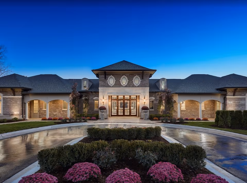 Front entrance of a large brick building with a circular driveway, manicured hedges and pink flowers under a deep blue twilight sky.