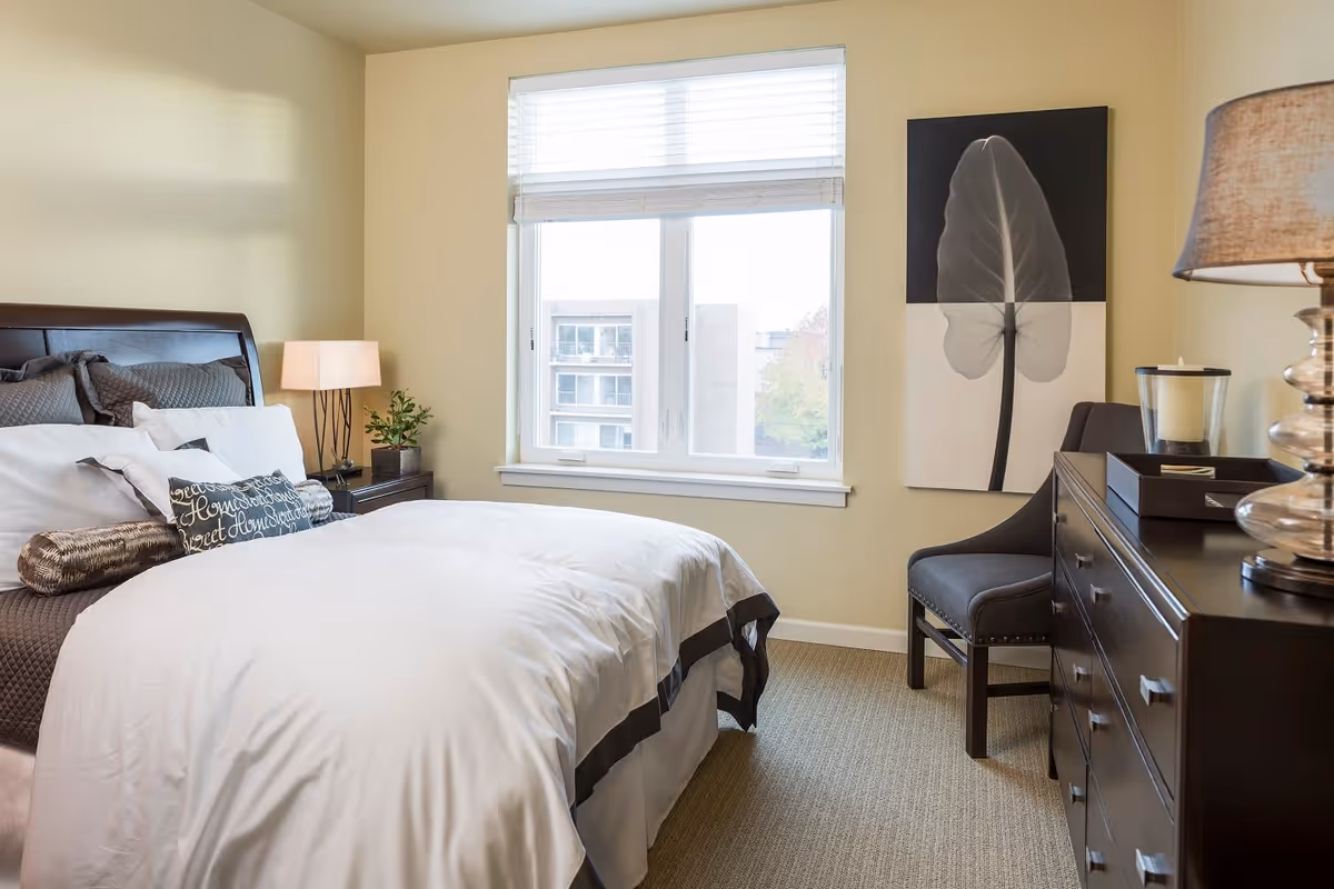 A neatly made bed with white and gray bedding in a bedroom with beige walls. There is a window with blinds letting in natural light, a nightstand with a lamp and a small plant, a dark wooden dresser with a lamp and candle, a gray upholstered chair, and a large black and white leaf artwork on the wall.