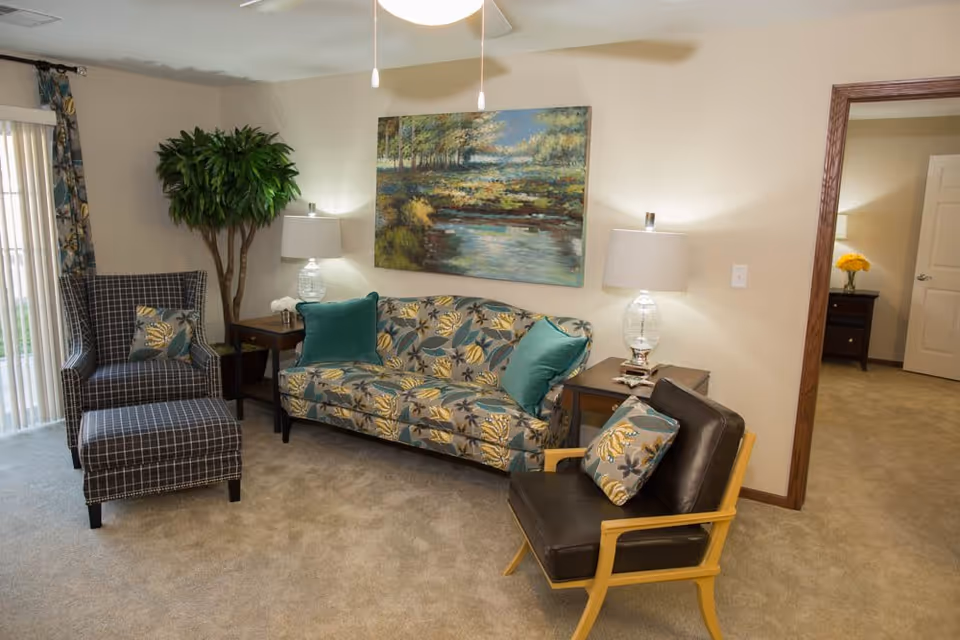 A cozy living room with a patterned sofa adorned with teal cushions, a black leather chair with a matching floral cushion, and a checkered armchair with an ottoman. Two side tables with glass lamps flank the sofa, and a large painting of a serene landscape hangs on the wall above it. A tall green plant is placed in the corner near sliding glass doors with vertical blinds. An open doorway leads to another room with a nightstand and a vase of yellow flowers.