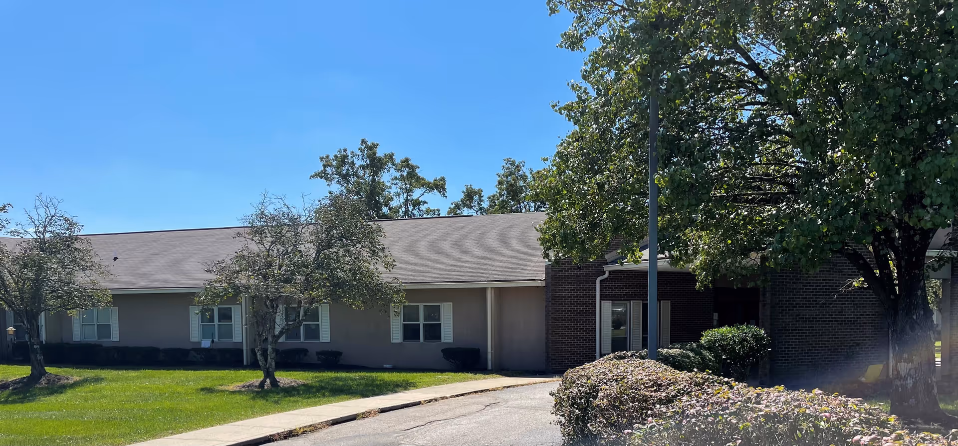 Single-story healthcare facility building frontage with windows, trees, a driveway and landscaped lawn under a clear blue sky.