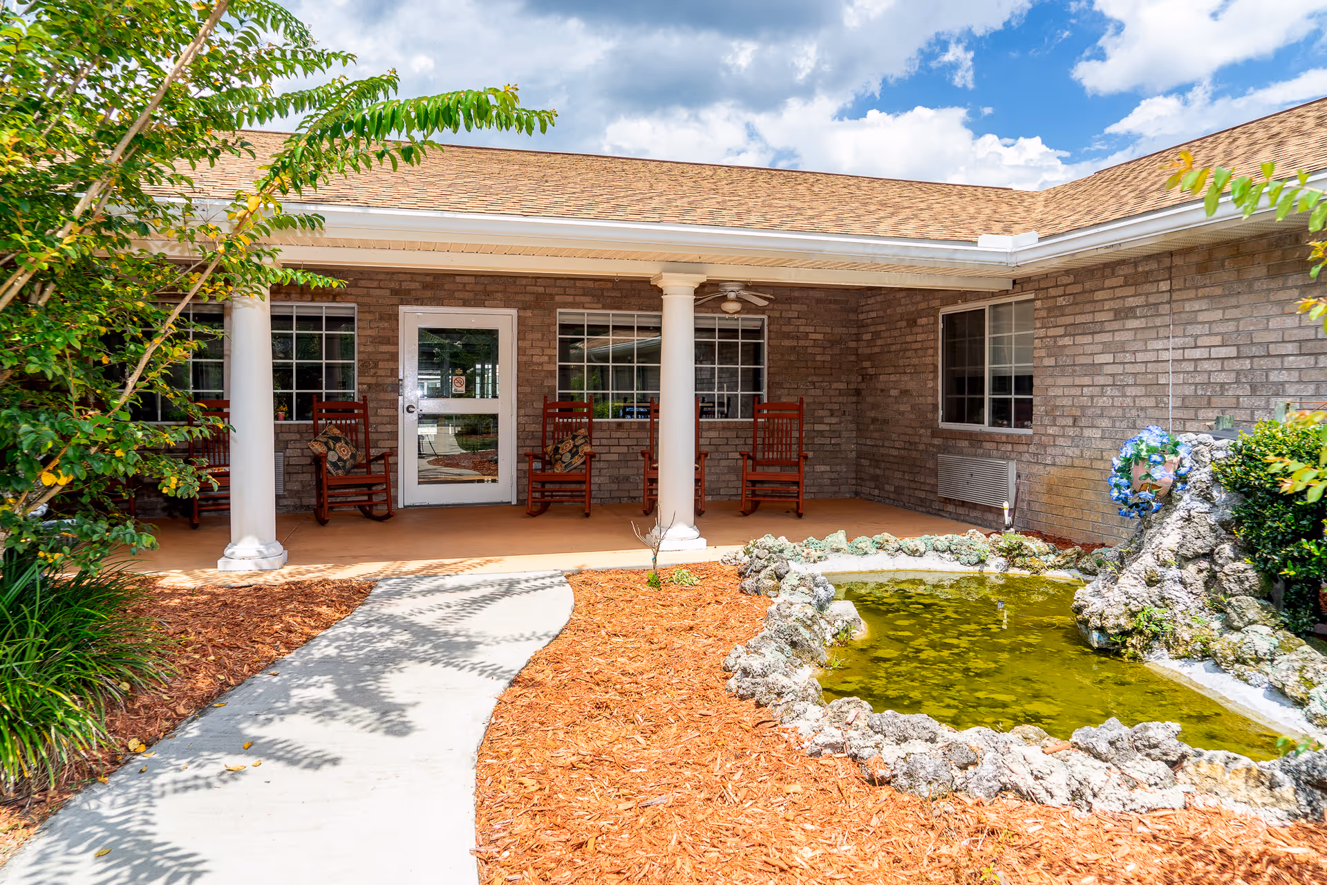 Covered brick entrance to a nursing home with a walkway, white columns, rocking chairs on a porch and a small decorative pond.