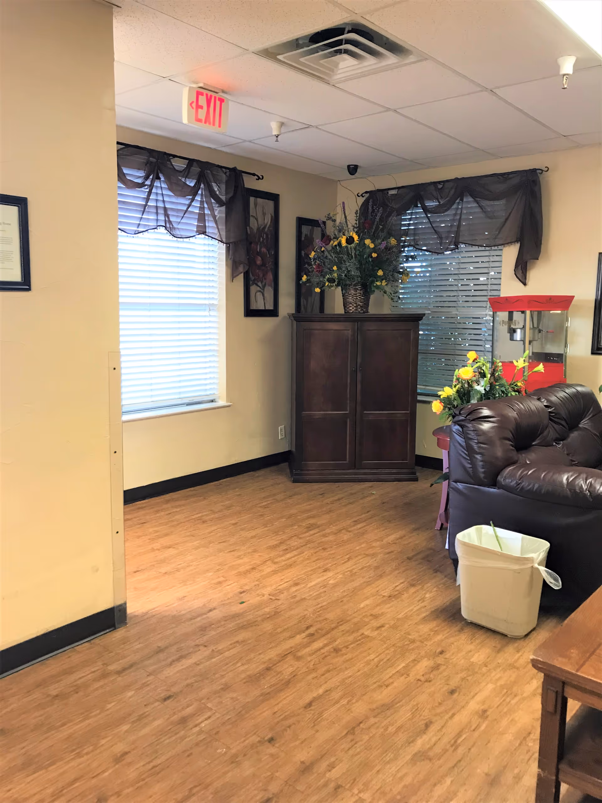 Interior corner of a room with wooden flooring, beige walls, and two windows with dark sheer curtains. A dark wooden cabinet with a large floral arrangement on top is positioned between the windows. To the right, there is a dark brown leather couch with a small table holding a yellow flower arrangement and a red popcorn machine behind it. A white trash bin is placed on the floor near the couch. An exit sign is visible on the ceiling near the corner.