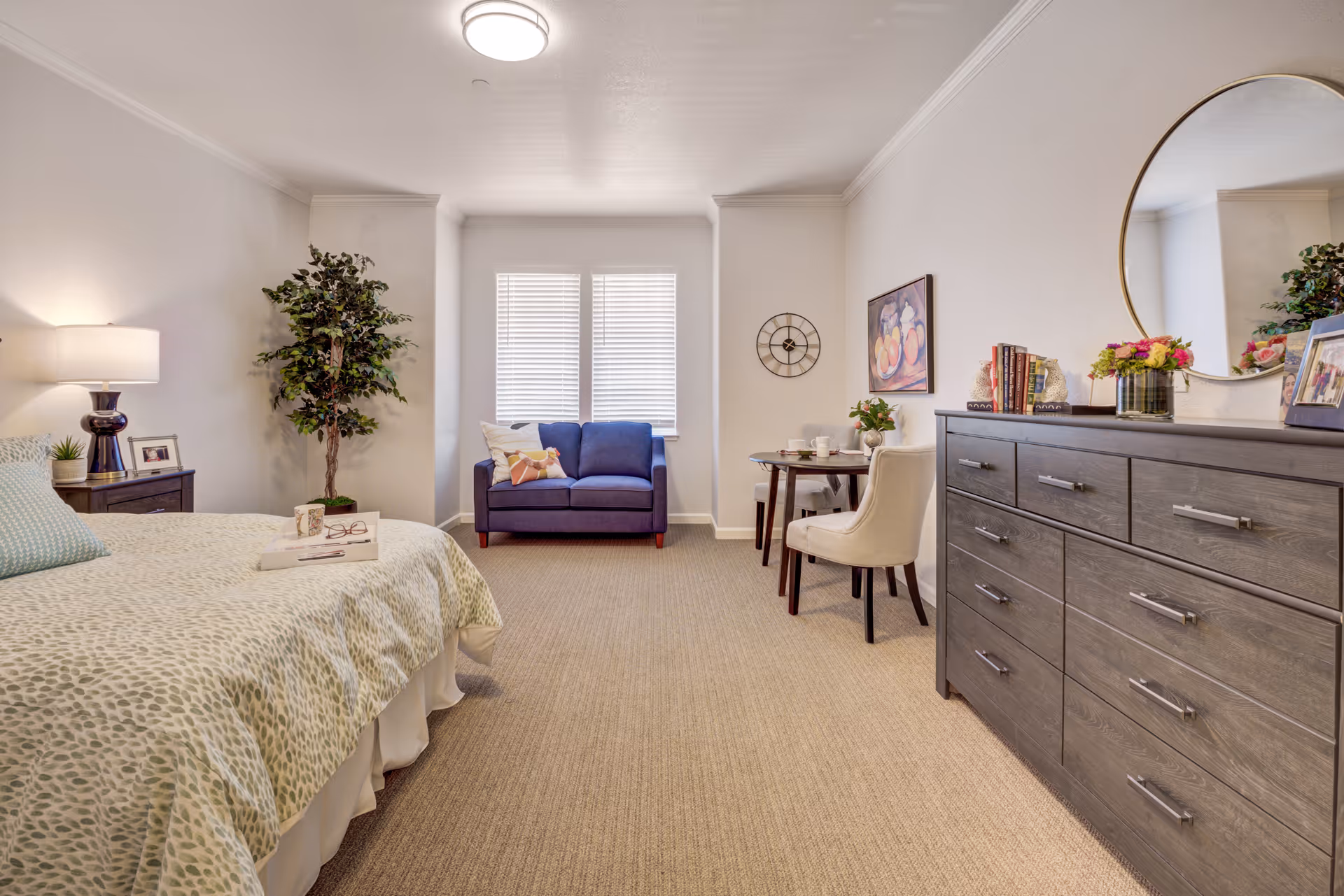 A bright and cozy senior living bedroom featuring a bed with patterned bedding, a bedside table with a lamp and picture frame, a potted plant, a blue loveseat under two windows with blinds, a small round dining table with two chairs, a wall clock, a painting, and a large dresser with a round mirror, books, flowers, and framed photos.