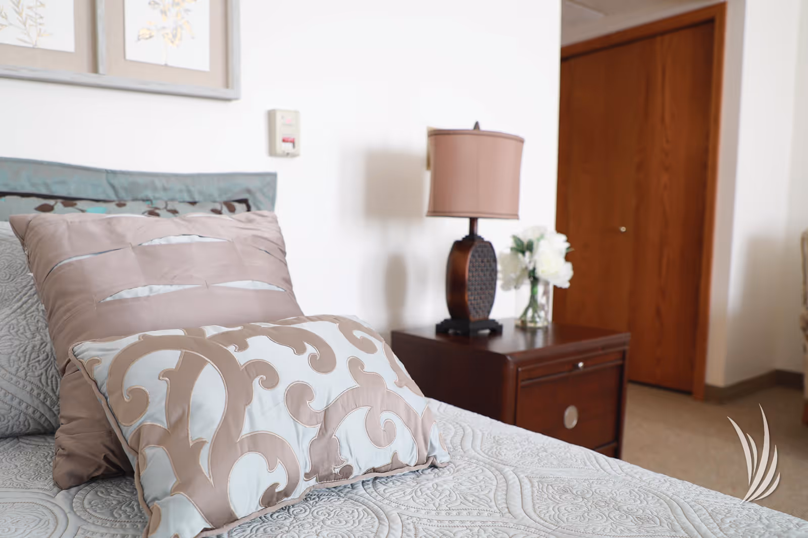 Close-up view of a bed with decorative pillows and a patterned bedspread in a bedroom. Next to the bed is a wooden nightstand with a lamp and a vase of white flowers. A wooden door is visible in the background.